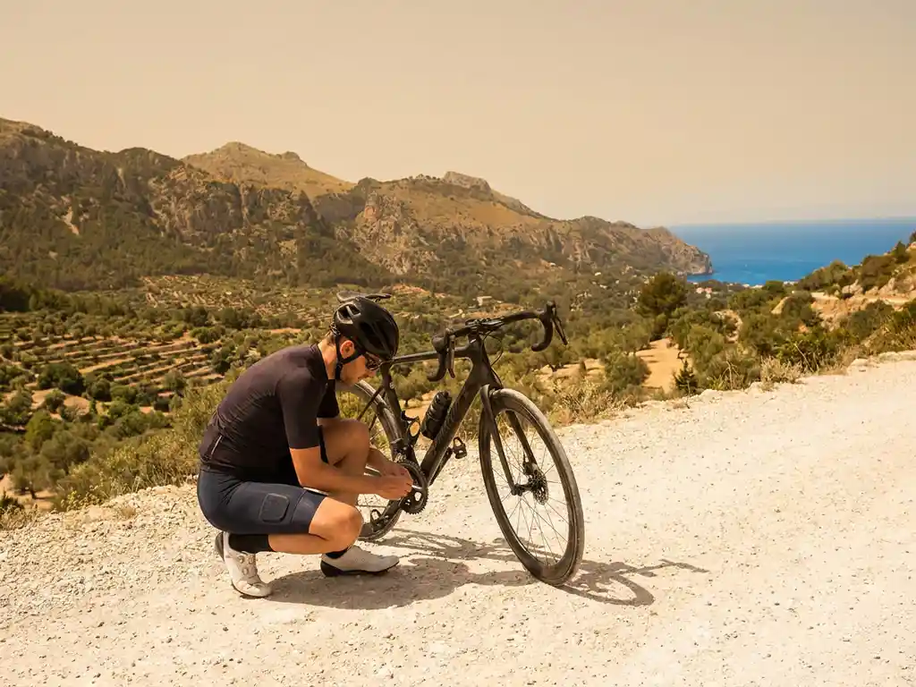 Cyclist adjusting padded cycling shorts beside a racing bike on a limestone road in Mallorca, with Serra de Tramuntana mountains and Mediterranean sea behind.
