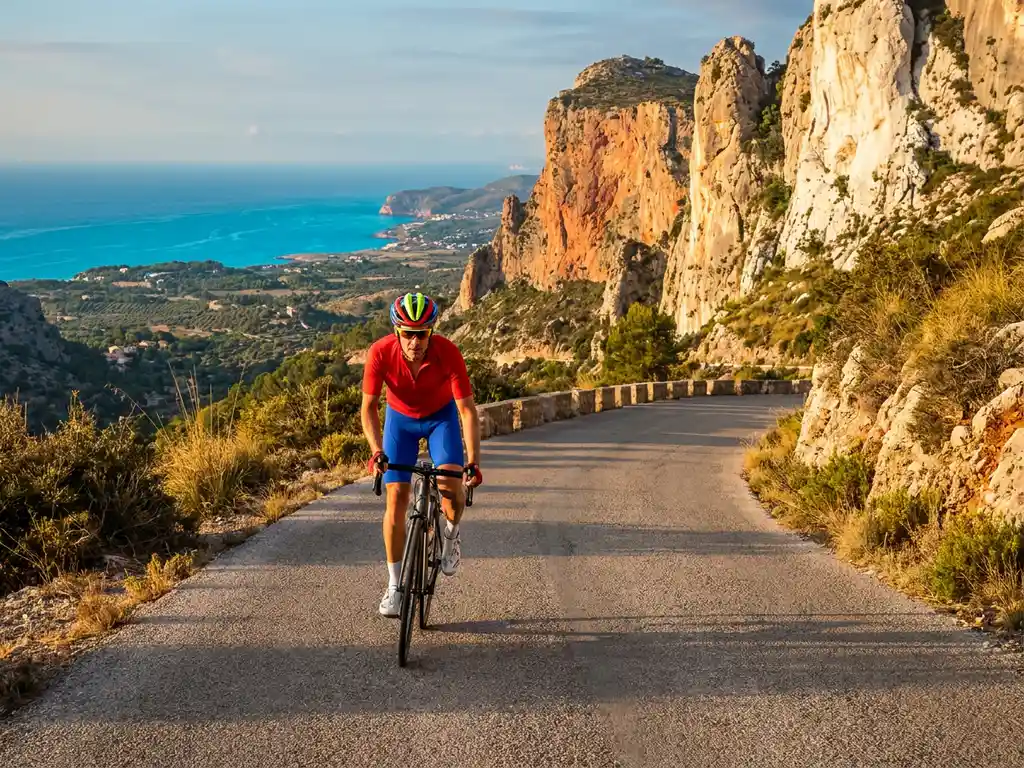 Cyclist in red kit cresting a Mallorca mountain pass, Serra de Tramuntana limestone cliffs and turquoise Mediterranean sea in golden afternoon light.