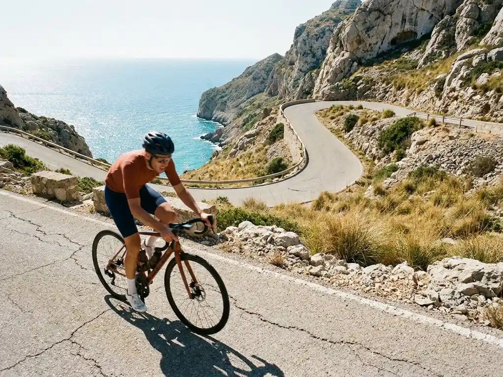 Radsportler erklimmt serpentinenreiche Bergstraße in Mallorcas Kalksteinklippen mit türkisblauem Mittelmeer im Hintergrund.