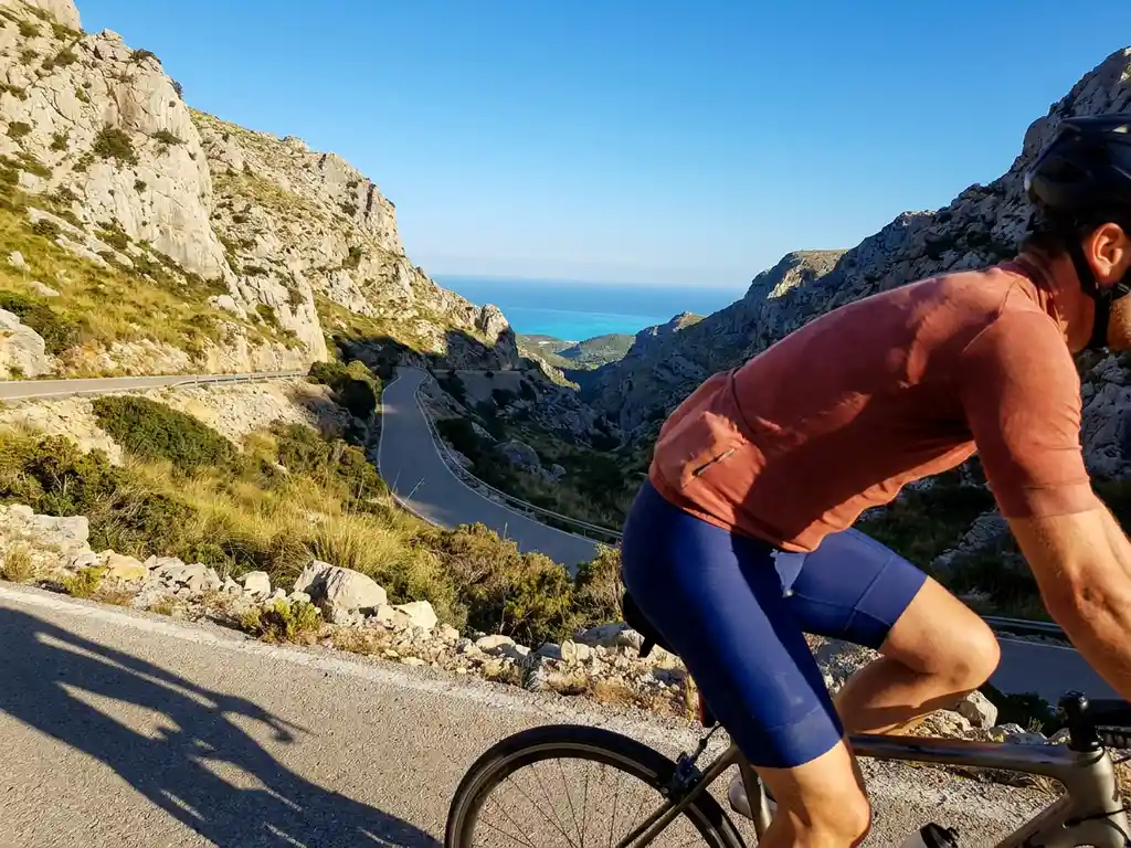 Wielrenner bereikt bergpas in Mallorca met kalkstenen kliffen van Serra de Tramuntana en turquoise Middellandse Zee op de achtergrond.